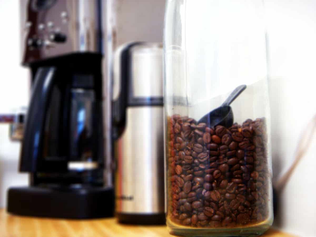Coffee beans in a jar with a grinder and coffee pot in background.