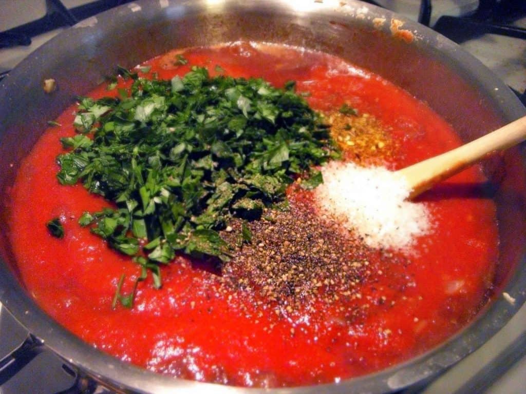 Marinara sauce in a cooking pot, with chopped basil, salt, and pepper about to be stirred in by a wooden spoon resting on the side of the pot.