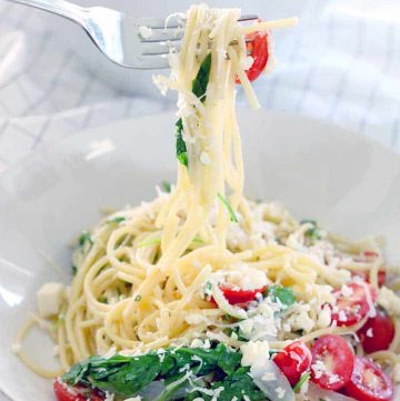 Fork twirling pasta with tomatoes, greens, and cheese, and lifted over a white plate.