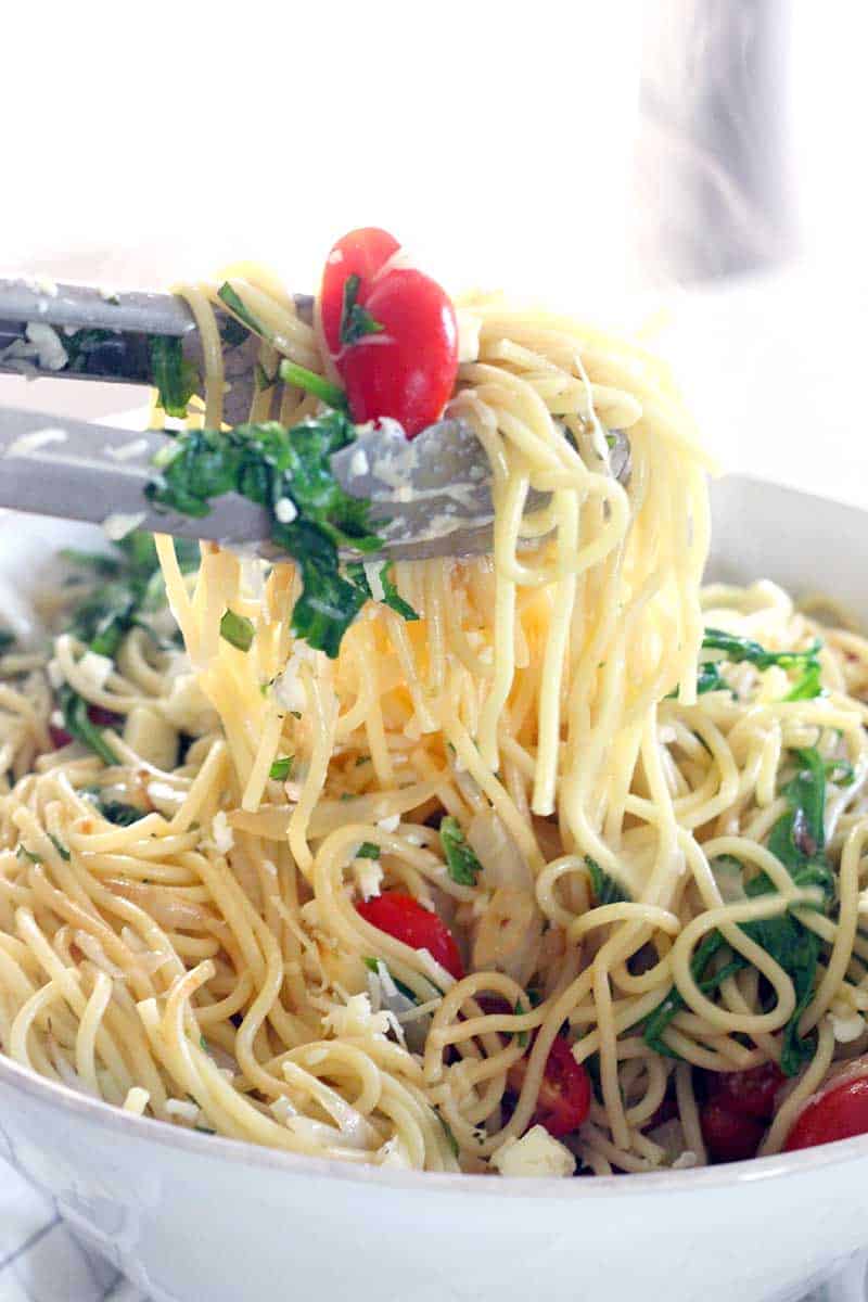 Tongs lifting pasta with arugula, tomatoes, and cheese out of a white bowl.