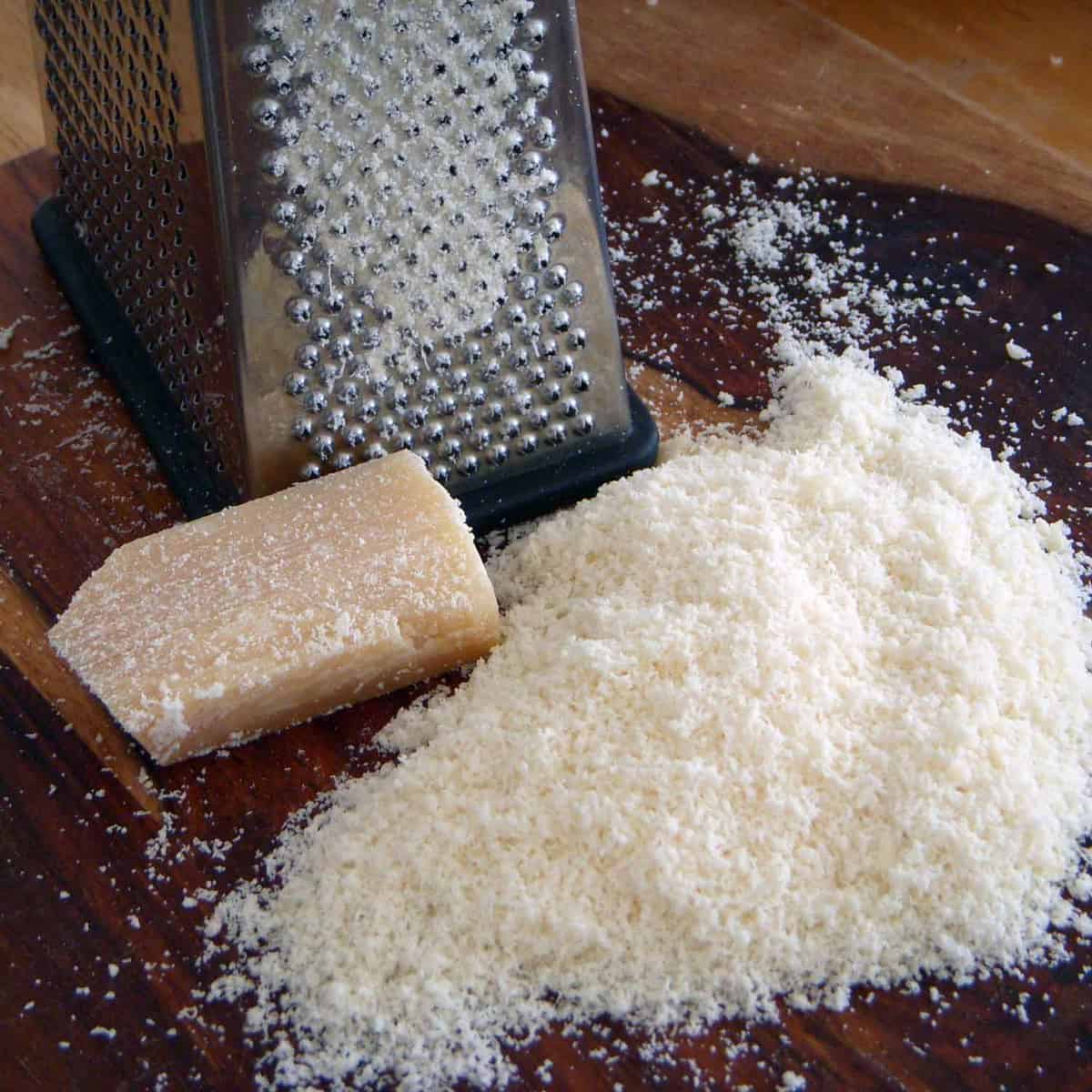 Grated and solid parmesan cheese and a grater on a wooden cutting board.