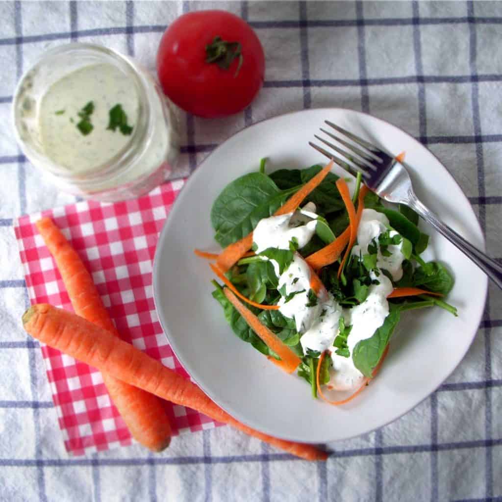 Bird's eye view of salad topped with ranch dressing on a white plate, surrounded by fresh produce and glass jar filled with ranch dressing.