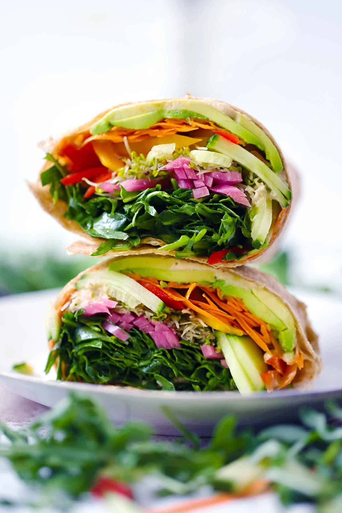 A veggie wrap cut in half and stacked on a white plate, showing the rainbow of vegetables, hummus, and avocado inside.