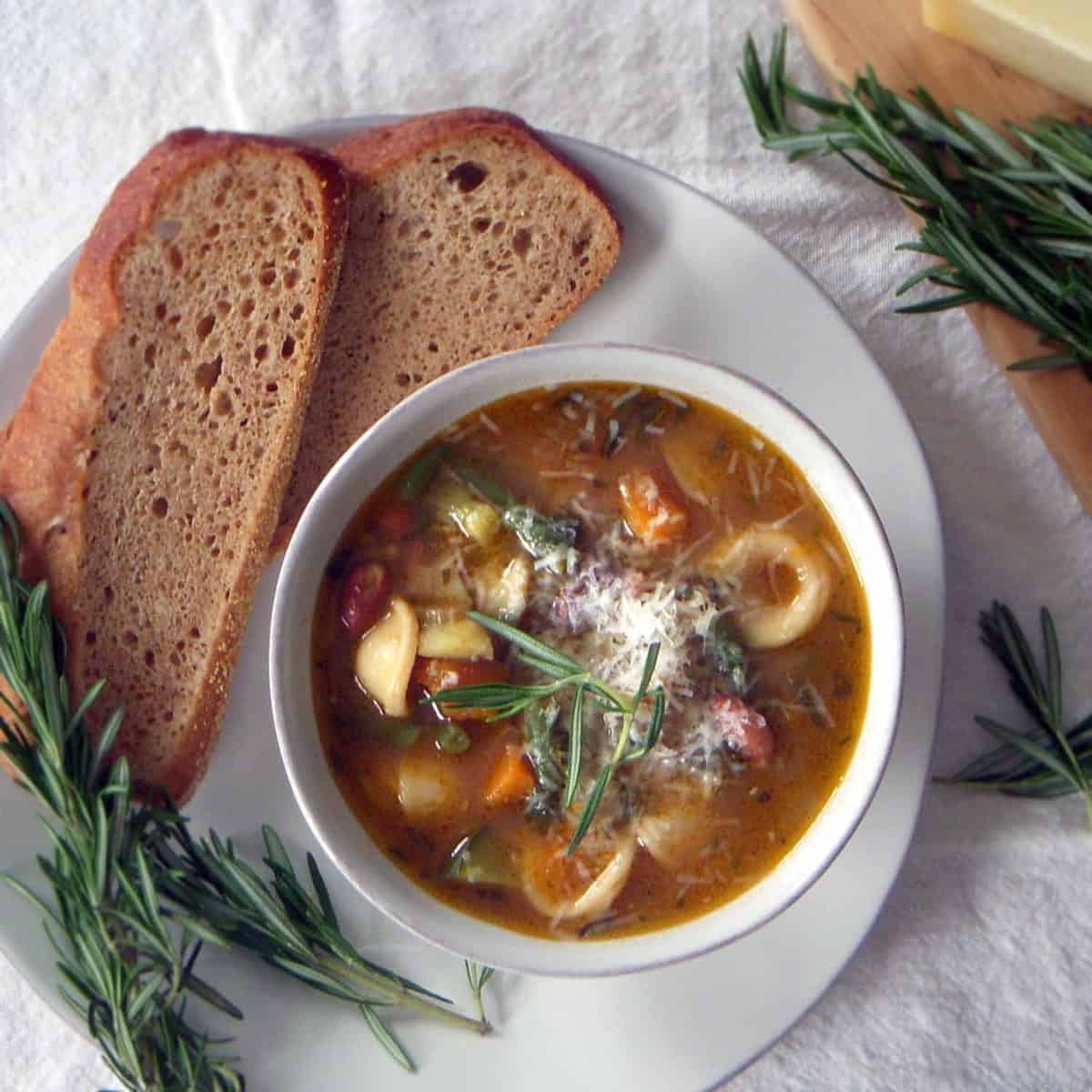 Bird's eye view of minestrone soup in a white bowl, on a white plate, with bread slices and a sprig of rosemary on the side.