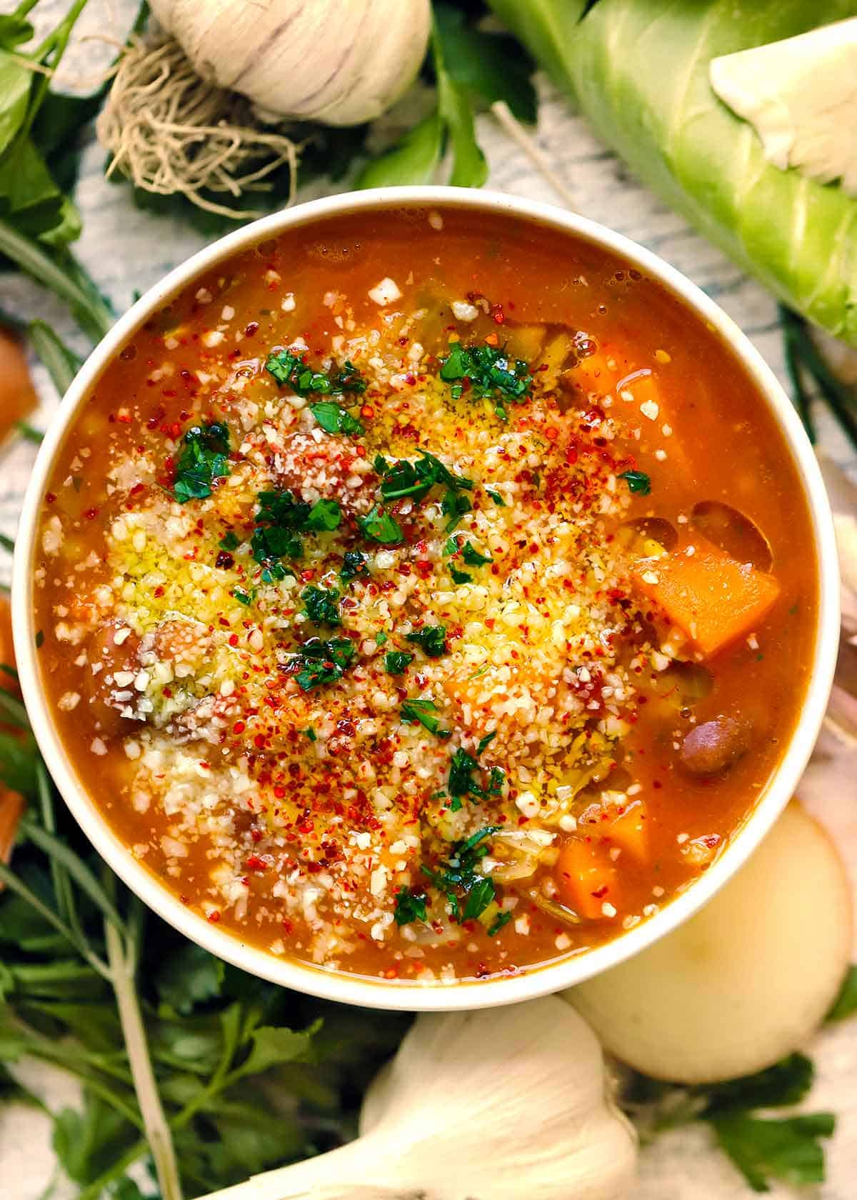 Overhead view of autumn harvest minestrone soup topped with grated cheese, red pepper flakes, and parsley in a white bowl, surrounded by garlic bulbs, cabbage, and fresh herbs.