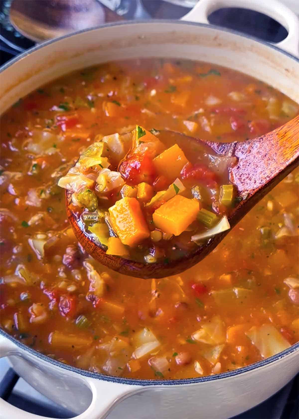 Wooden ladle lifting a scoop of autumn minestrone soup with butternut squash, cabbage, celery, and tomatoes from a cream-colored Dutch oven.