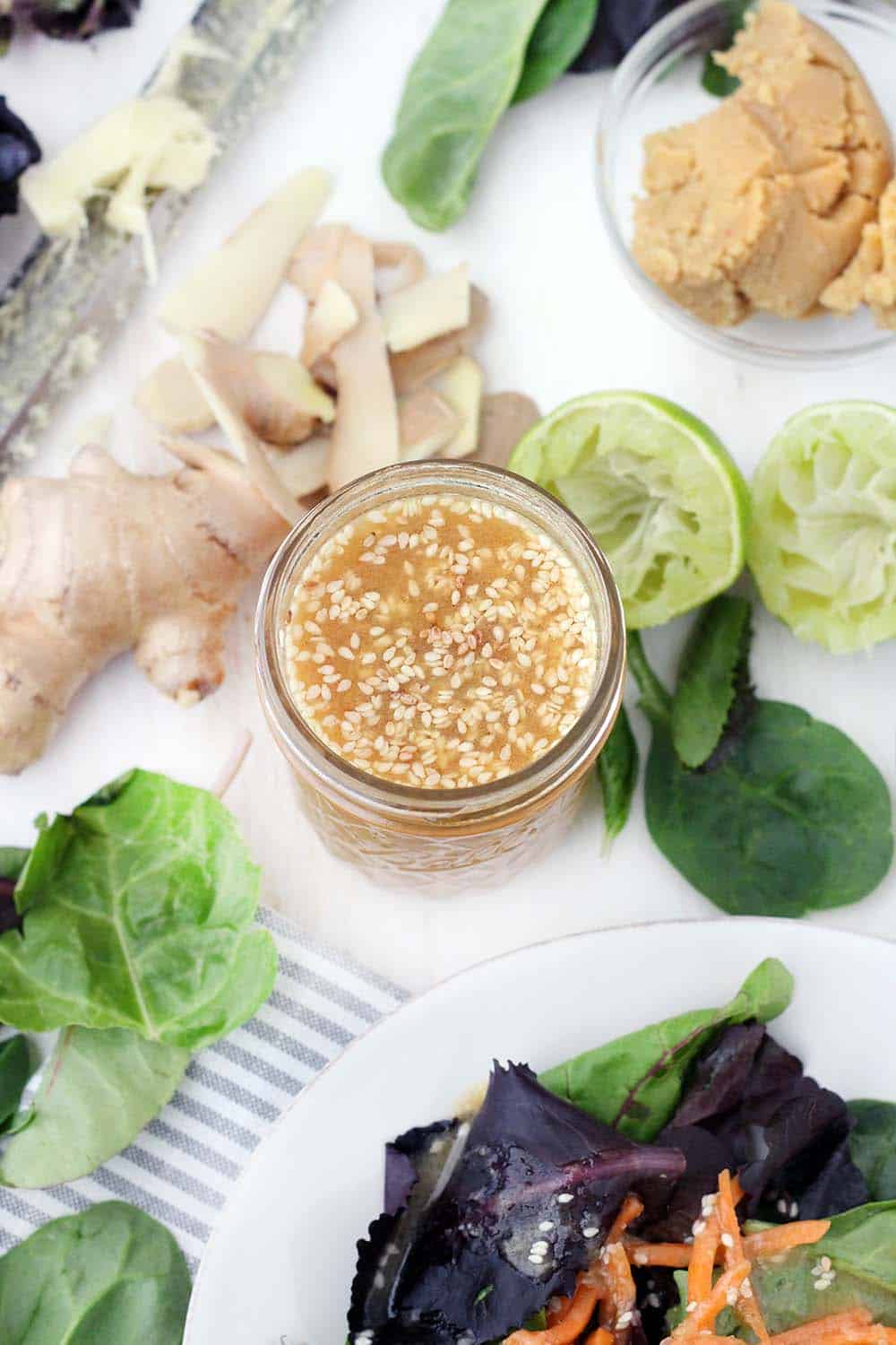 Bird's eye view of open glass jar filled with peanut dressing with sesame seeds mixed in, on white surface with fresh produce scattered around the jar.