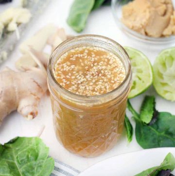 Open glass jar filled with peanut dressing with sesame seeds mixed in, on white surface with fresh produce scattered around the jar.
