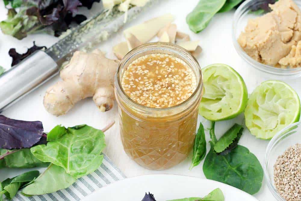 Horizontal photo of open glass jar of peanut dressing, with lettuce and other fresh produce scattered around the jar.
