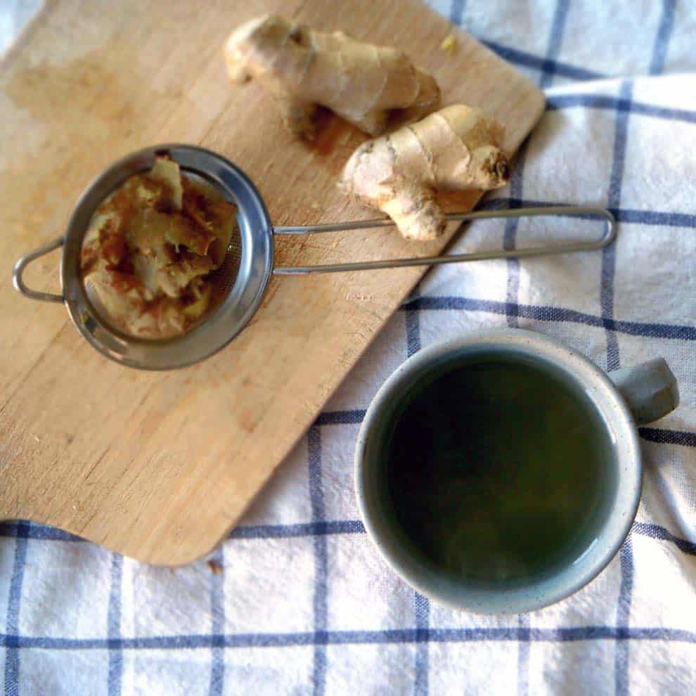 Bird's eye view of tea in a ceramic mig, on a blue and white checkered cloth with a wooden cutting board, a small mesh strainer, and ginger root on the side.