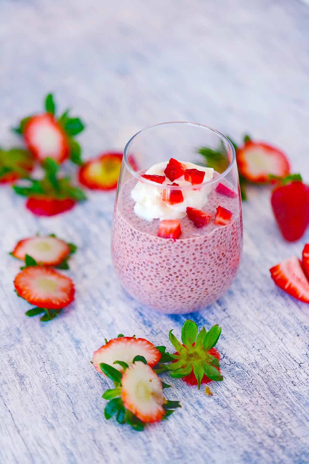 A glass filled with strawberry chia seed pudding with fresh strawberry scraps scattered around, topped with fresh strawberries and whipped cream.