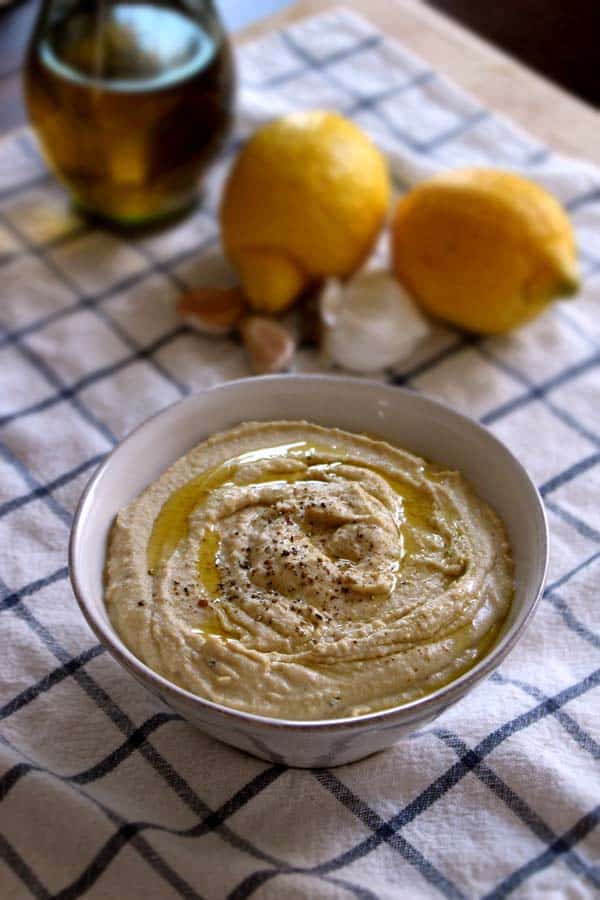 Bowl of hummus in foreground on a blue and white checkered cloth, with lemons and bottle of olive oil in background.
