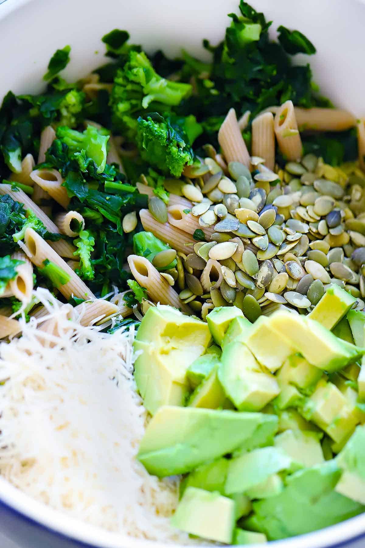 Mixing up whole wheat pasta, broccoli, spinach, avocado, pumpkin seeds, and parmesan cheese in a large bowl.