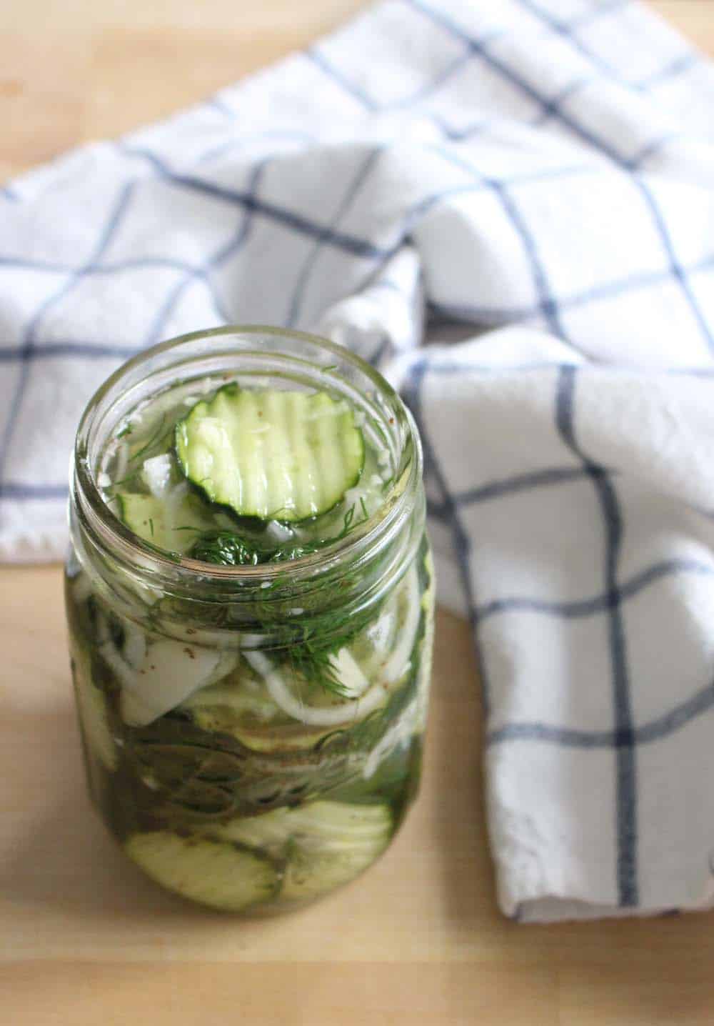 Open glass jar filled with pickles in their juice with herbs, on a wooden table next to a blue and white checkered cloth.