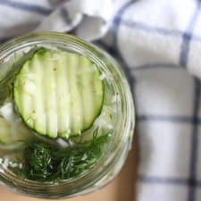 Bird's eye view of an open glass jar filled with pickles in their juice and fresh herbs, on a wooden table, next to a blue and white checkered cloth.