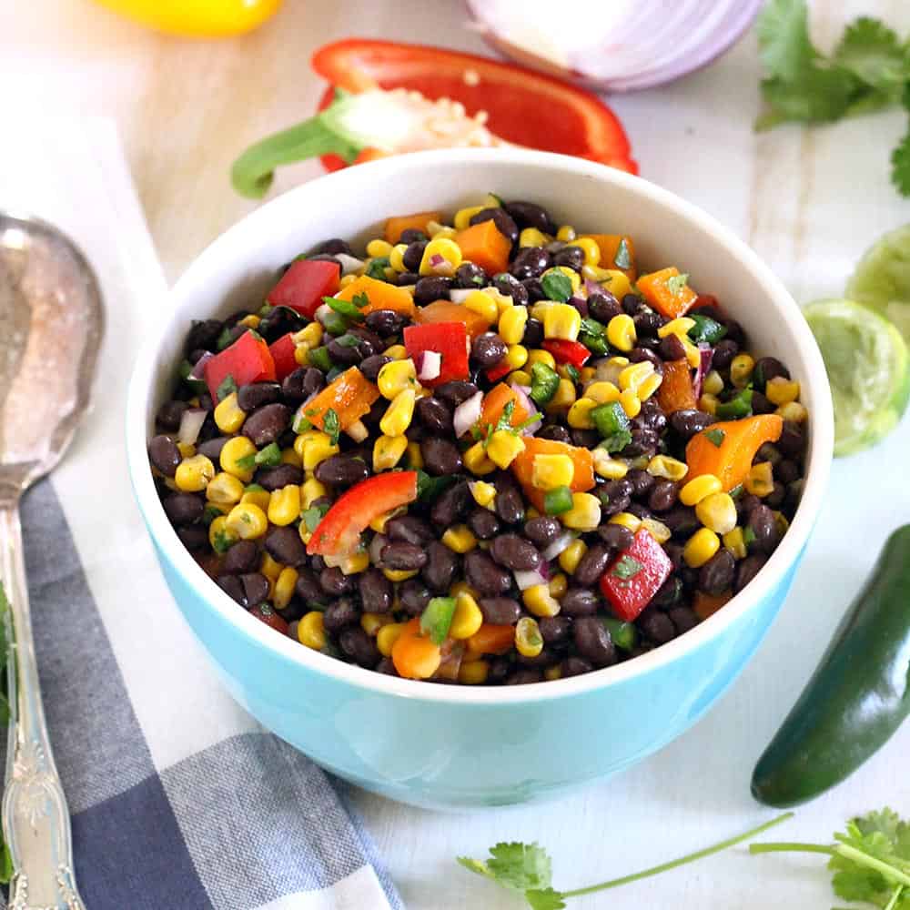 Black bean and corn salad in a light blue bowl, on a white surface with produce scattered around.