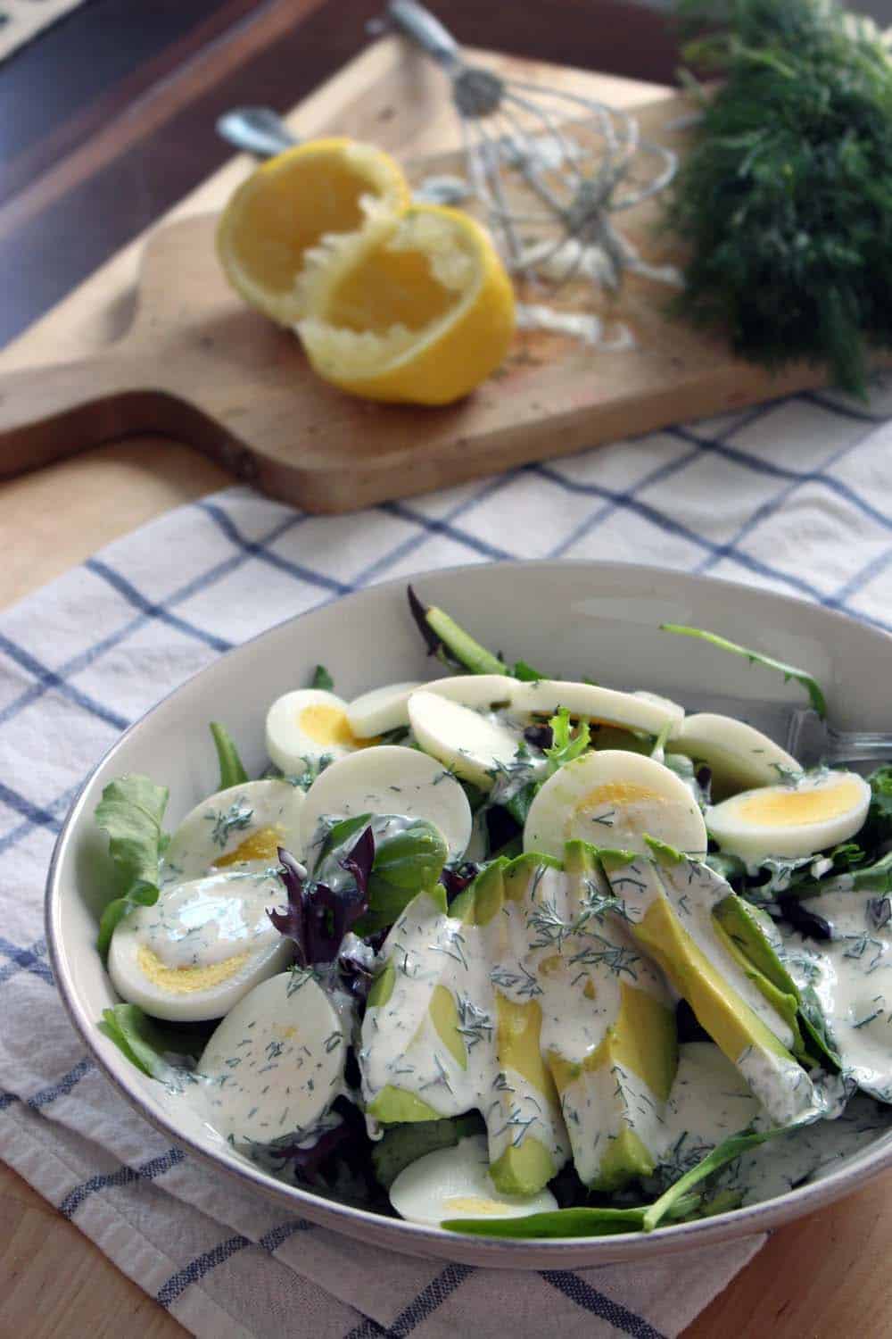 Mixed greens salad with egg and avocado, topped with creamy white dressing, in a white bowl on a blue and white checkered cloth. A wood cutting board with lemons and dill is in the background.