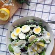 Bird's eye view of mixed greens salad topped with egg and avocado in a white bowl, on a blue checkered cloth.