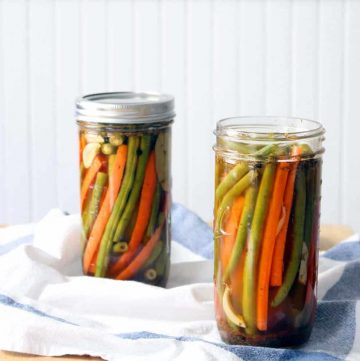 Two jars of pickled green beans and carrots; one on the left and in the background with the lid on, and one on the right with the lid off in the foreground.