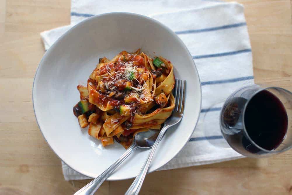 Bird's eye view of pappardelle pasta coated in red sauce in a white plate, with a fork and spoon, on a white and blue striped cloth with a glass of red wine on the side.