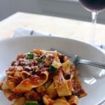 White plate holding pappardelle pasta coated with red sauce, being twirled with a fork. A glass of red wine is in the background.