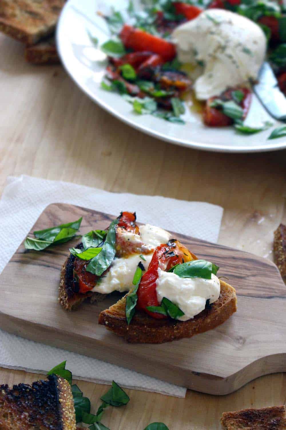 Small slice of toasted bread topped with burrata and roasted tomato, cut in half, on a wooden cutting board with a white plate holding recipe ingredients in the background.