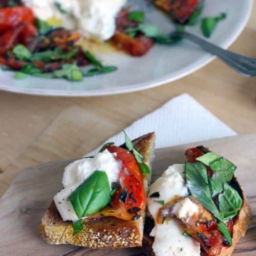 Halved roasted tomato and burrata toast on a wooden cutting board, with a white plate holding recipe ingredients in the background.
