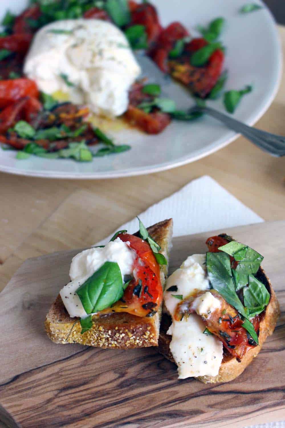 Halved roasted tomato and burrata toast on a wooden cutting board, with a white plate holding recipe ingredients in the background.