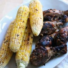 Four stacked ears of grilled corn on the cob next to grilled and seasoned chicken drumsticks on a white platter.