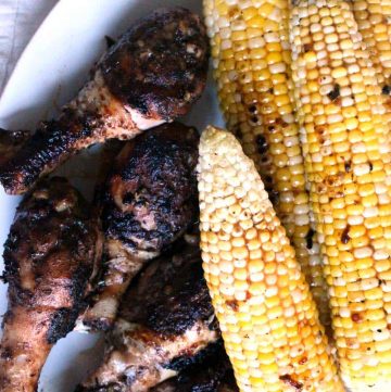 Bird's eye view of four grilled ears of corn on the cob next to several grilled and seasoned chicken drumsticks.