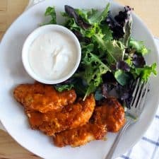 Bird's eye view of white plate holding buffalo chicken fingers, mixed greens, and a ramekin full of creamy white dipping sauce.