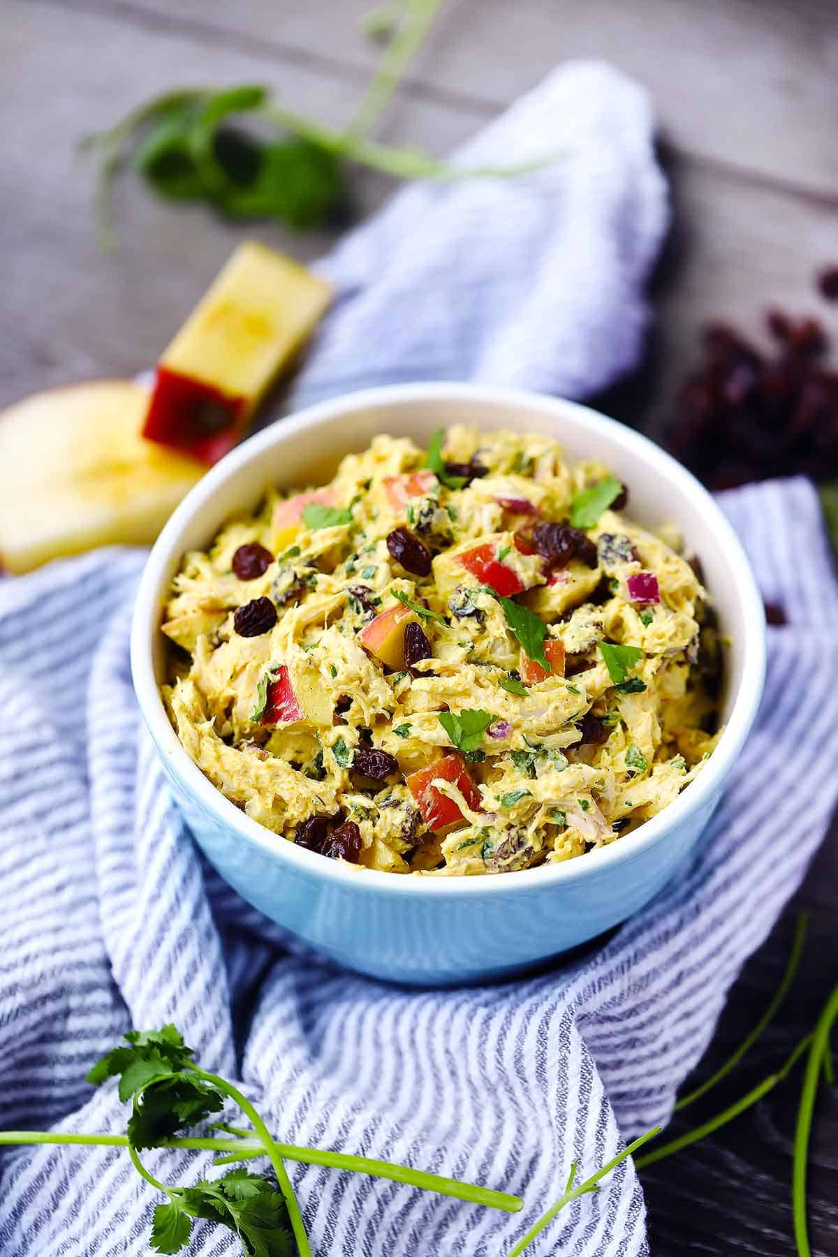 A bowl of curried chicken salad with apples and raisins in the background on a towel.