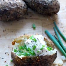 Baked potato sliced down the middle and topped with sour cream, pepper, and sliced green onions. Whole baked potatoes are in the background and whole scallions are displayed on the side.