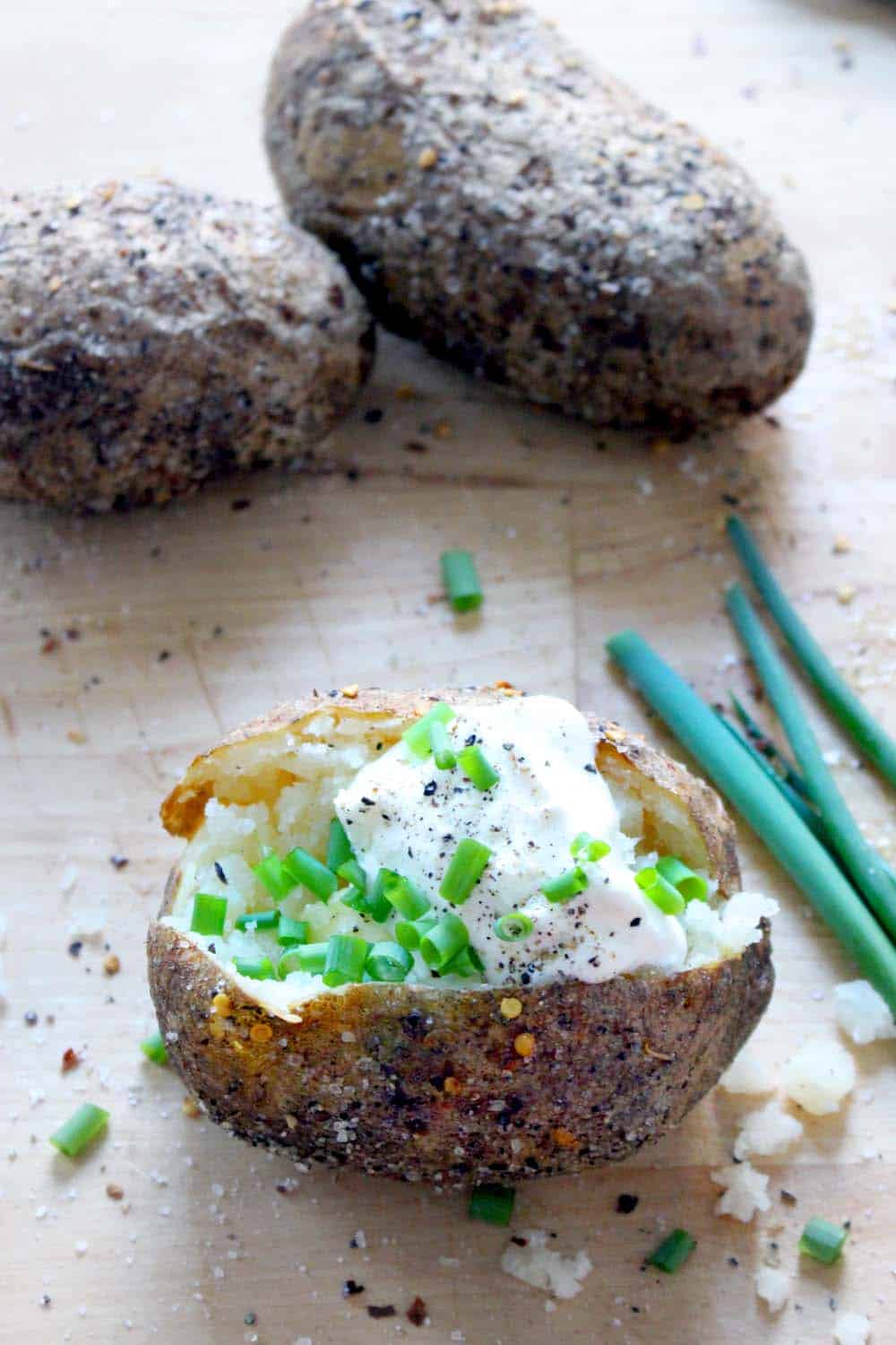 Baked potato sliced down the middle and topped with sour cream, pepper, and sliced green onions. Whole baked potatoes are in the background and whole scallions are displayed on the side.