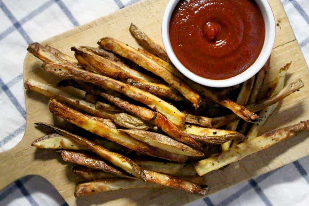 Bird's eye view of a pile of fries next to a ramekin of ketchup, all on a wooden cutting board.