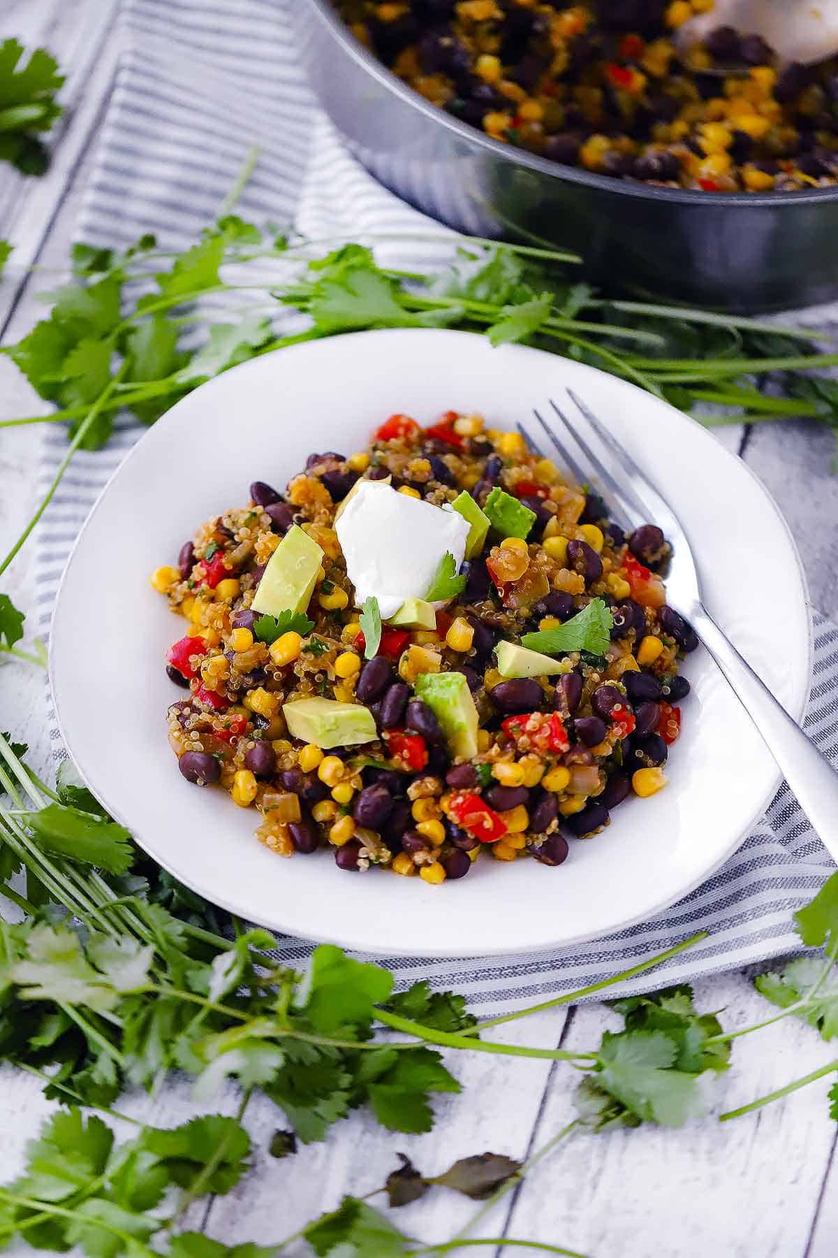 A white plate with black beans and quinoa topped with avocado and sour cream.