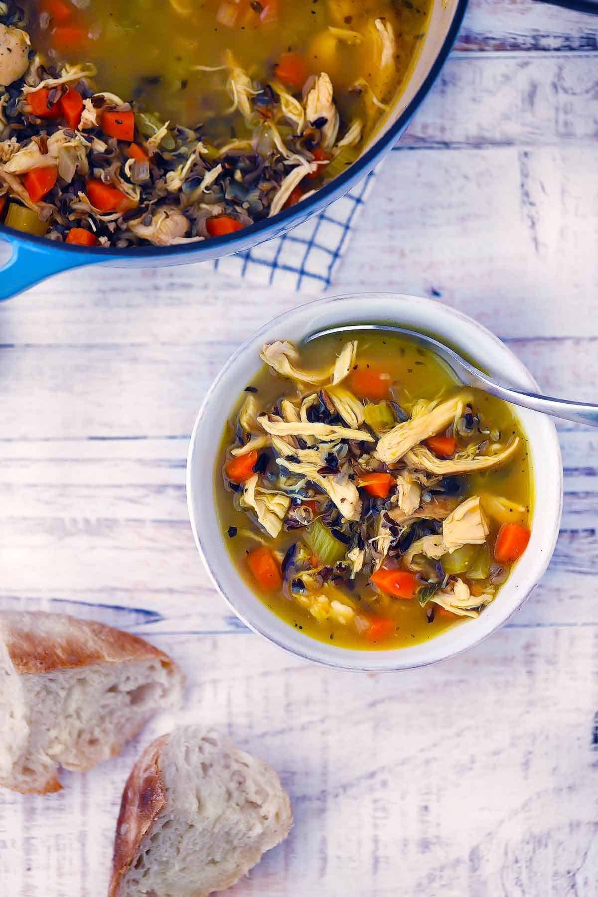 Bird's eye view of chicken wild rice soup in a white bowl with bread and a Dutch oven around it on a white wooden background.