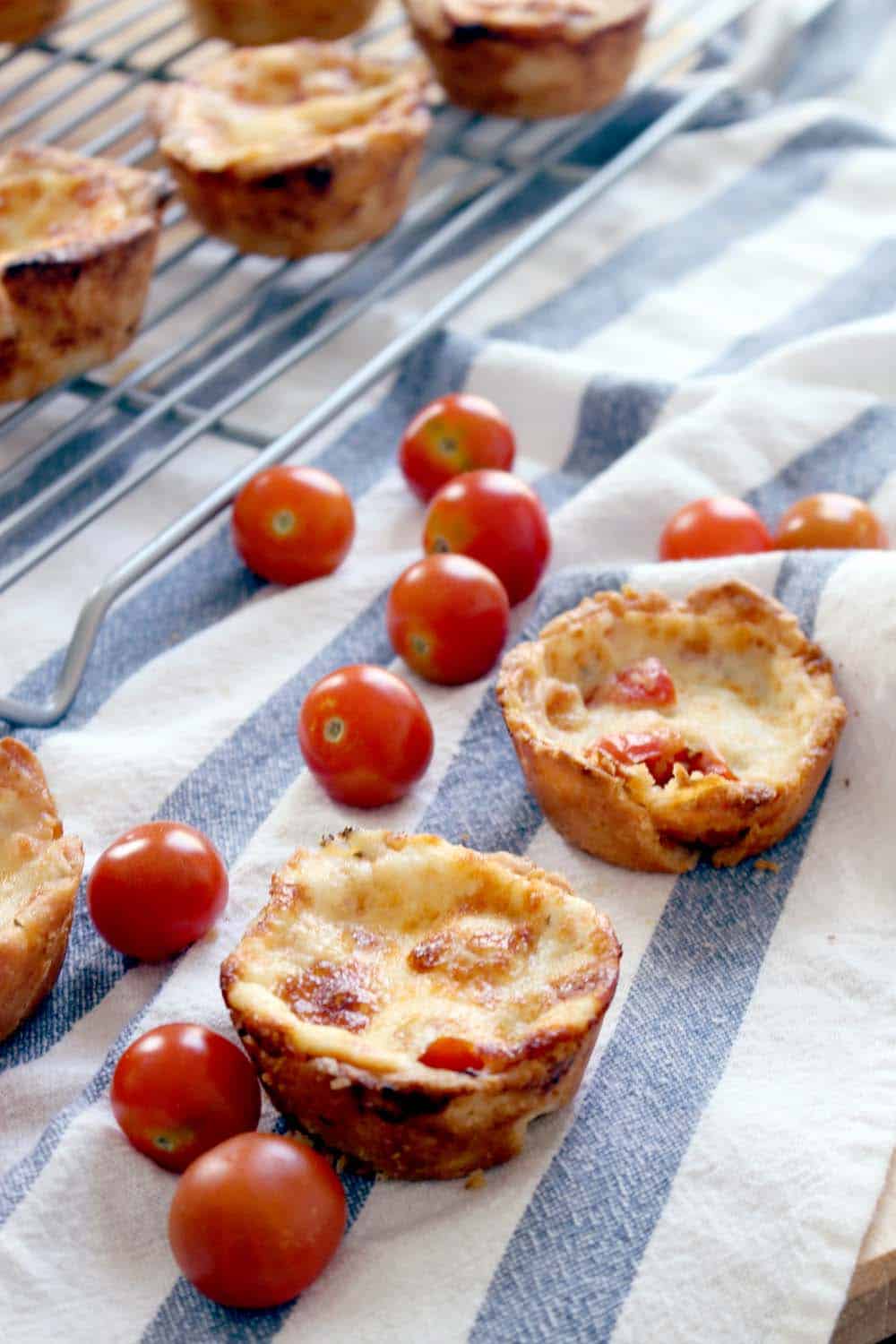 Two mini tomato pies in the foreground on a blue and white striped cloth with tomatoes scattered around them. A wooling rack holding more tomato pies is in the background.