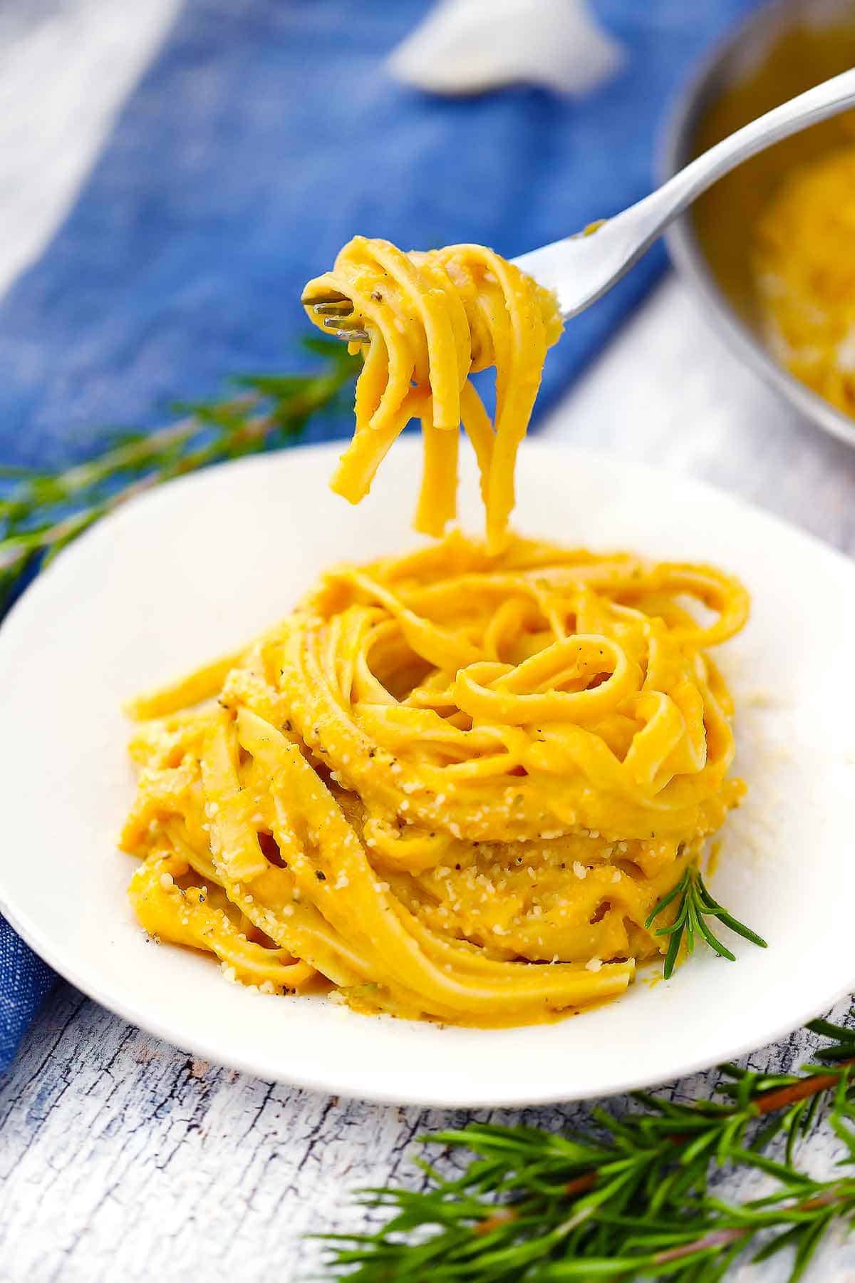 A white bowl holding butternut squash fettuccini, with a fork lifting some twirled pasta out of the bowl.