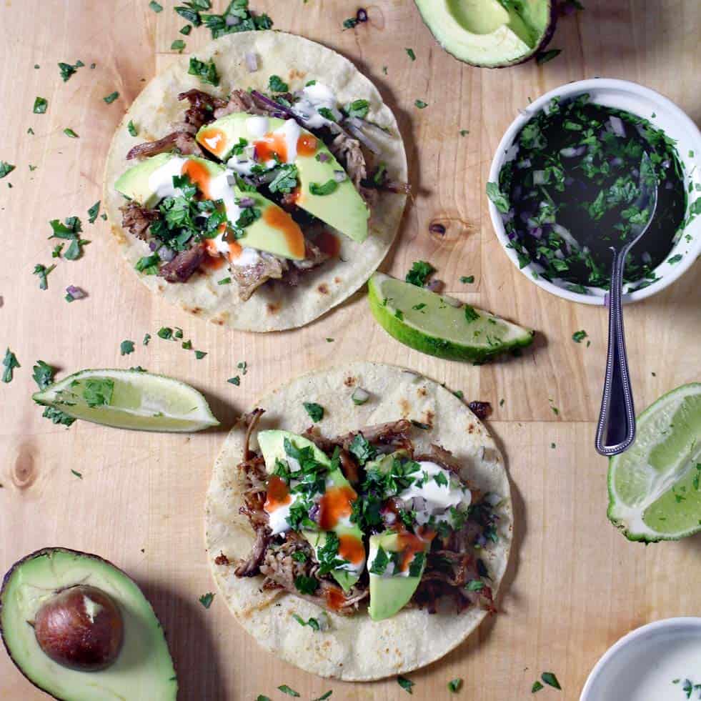 Bird's eye view of a wooden surface with open tortillas holding carnitas, sliced avocado, sour cream, hot sauce, and garnished with chopped herbs. Lime wedges, avocado, chopped herbs, and other ingredients are scattered around.