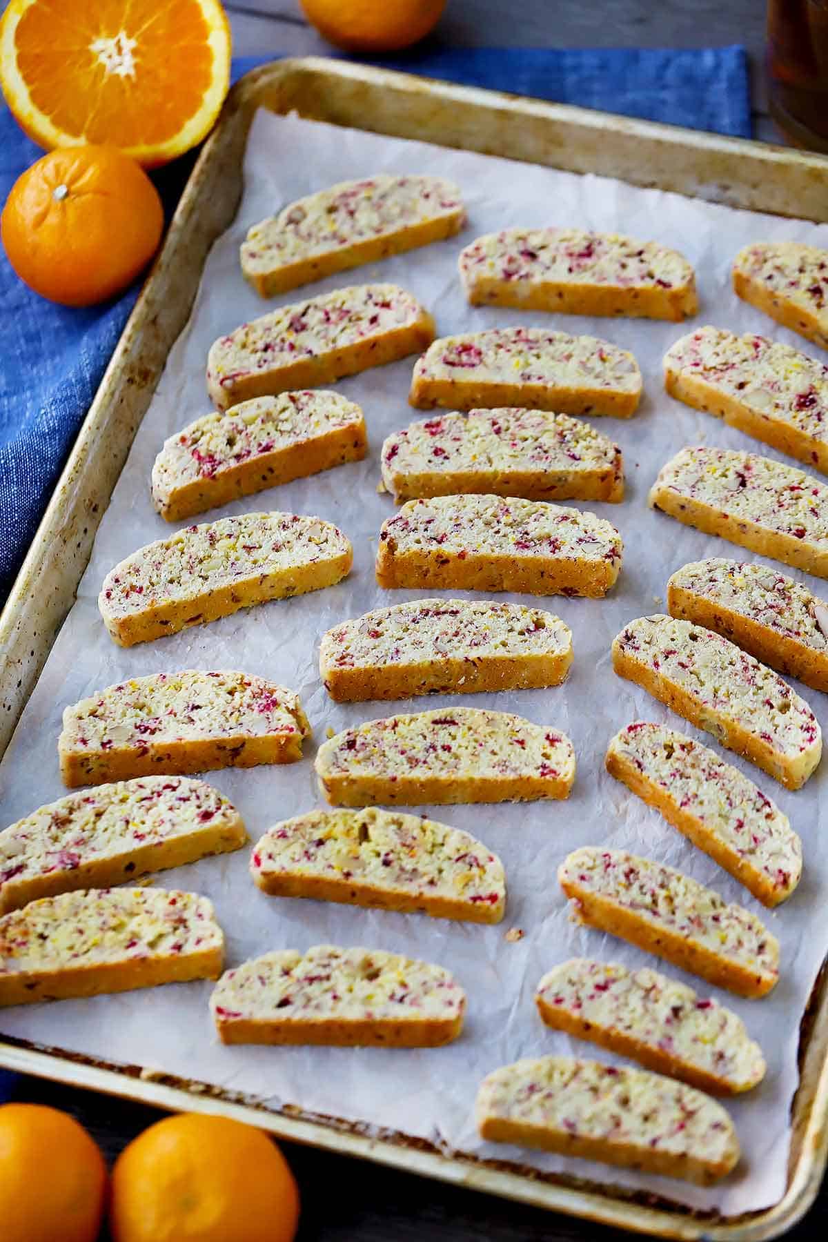 A baking sheet with biscotti on parchment paper with oranges in the background and a blue towel.