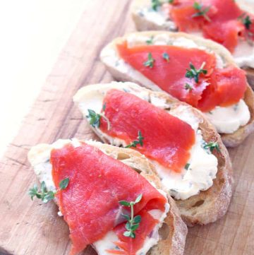 A row of four cream cheese and salmon toasts on a wooden cutting board.