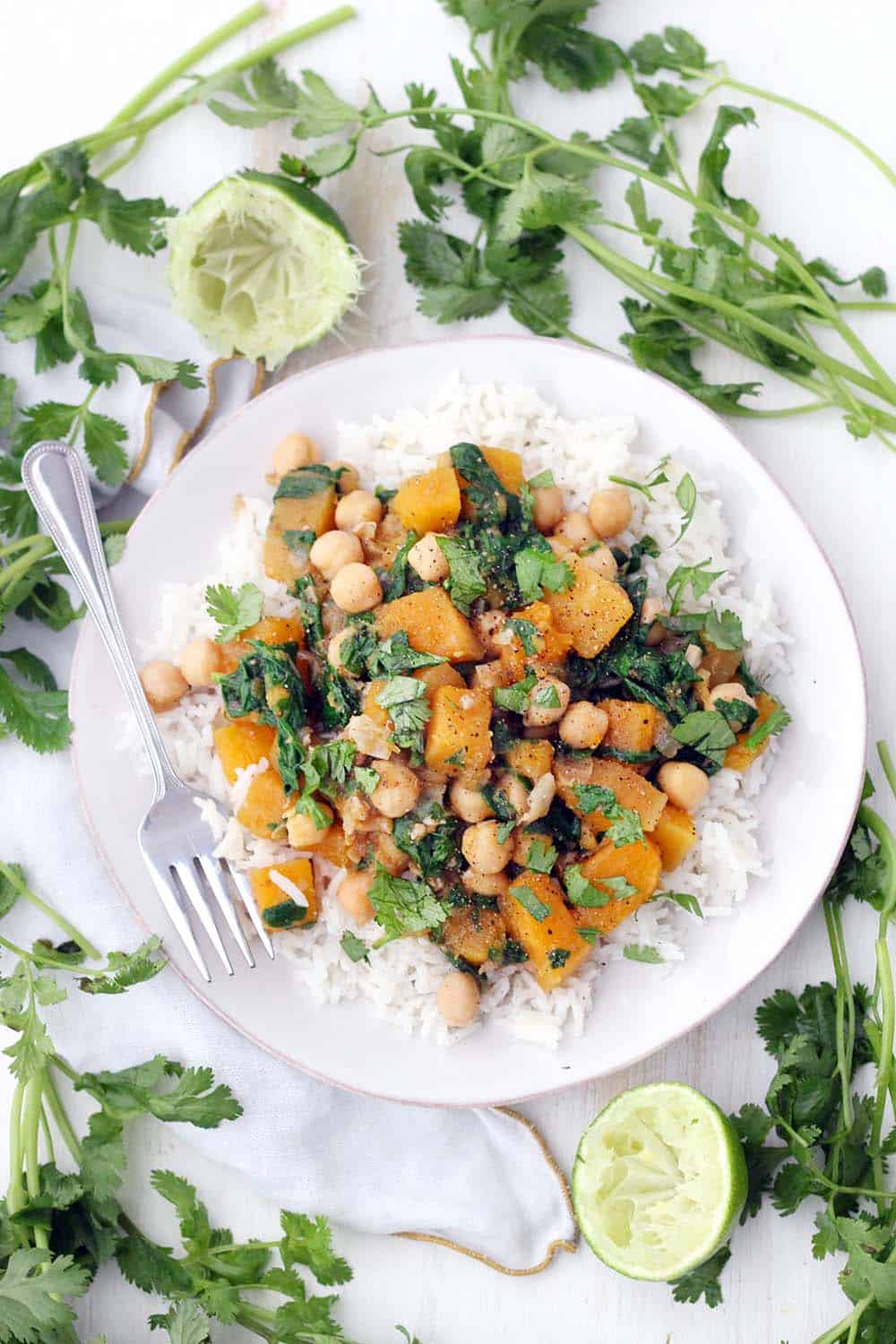 Bird's eye view of Moroccan butternut squash and chickpea stew on a bed of white rise, on a white plate, with a fork perched on the side and herbs scattered around.