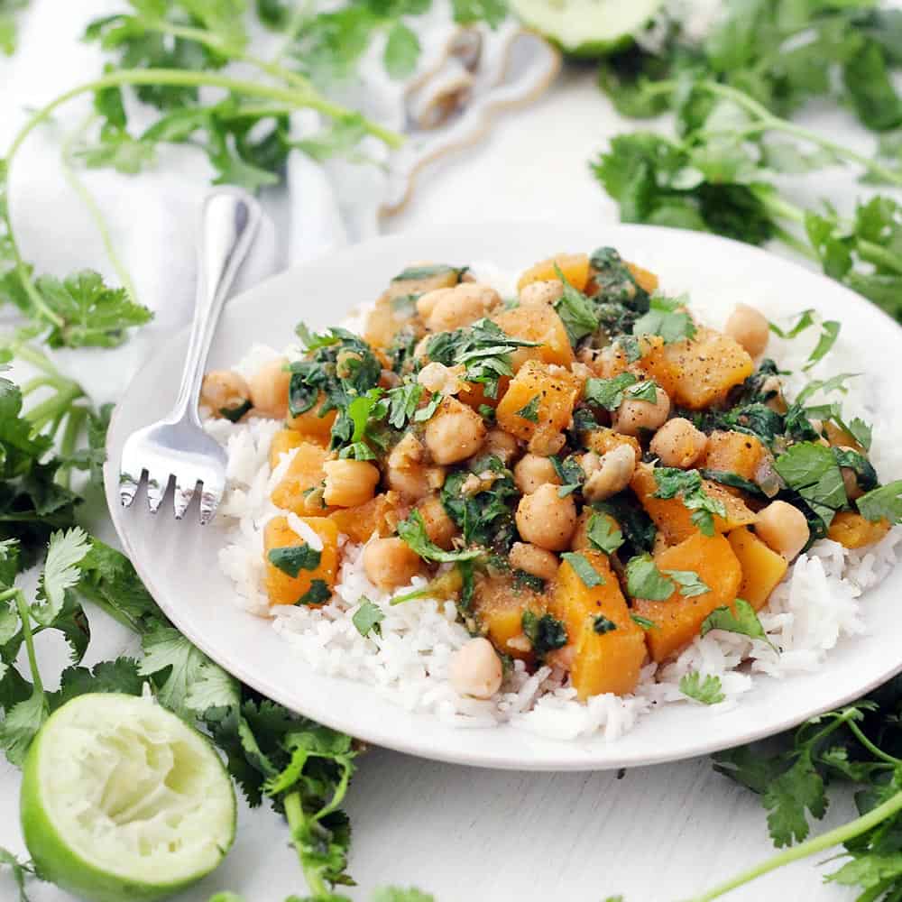 Side view of moroccan butternut squash and chickpea stew on a bed of white rise, on a white plate, with a fork perched on the side and herbs scattered around.