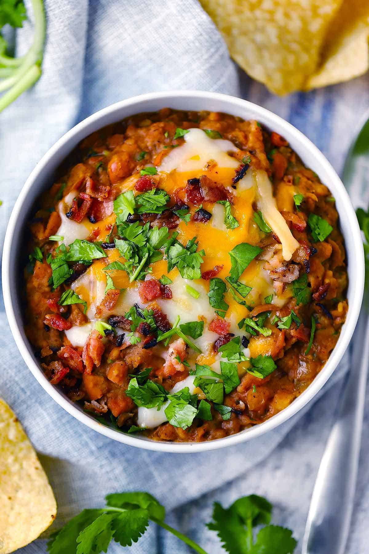 Overhead photo of a white bowl of refried beans with cheese, bacon, and cilantro on top.