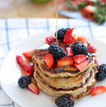 Stack of four pancakes on a white plate, topped with berries and a stream of maple syrup drizzling onto the pancakes from outside of the photo frame.