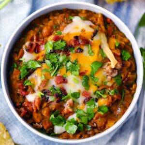 Square photo of refried beans in a white bowl with cheese, bacon, and cilantro on top.