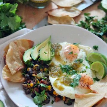 White plate holding fried eggs, tortilla chips, sliced avocado, and black beans, all garnished with fresh herbs.