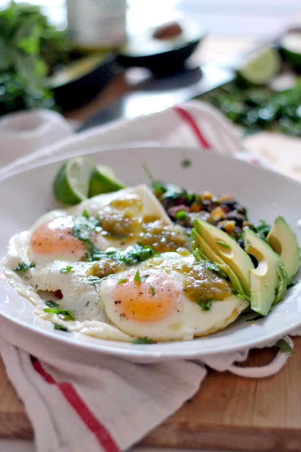 Two fried eggs on a plate with avocado slices, black beans, and chopped herb garnish on a white plate.