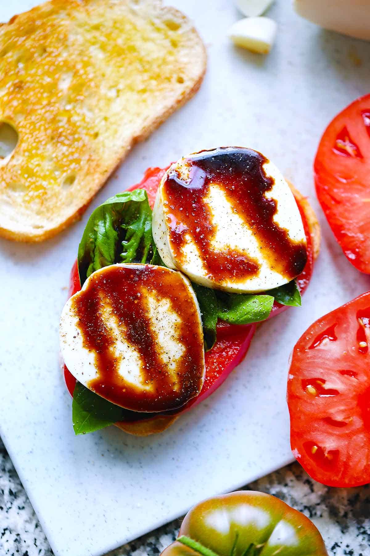 An open faced caprese sandwich with tomato, basil, and mozzarella, drizzled with balsamic glaze.